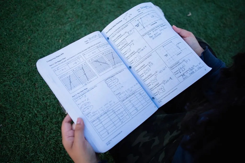 Open workbook with mathematical problems and graphs held over green grass.