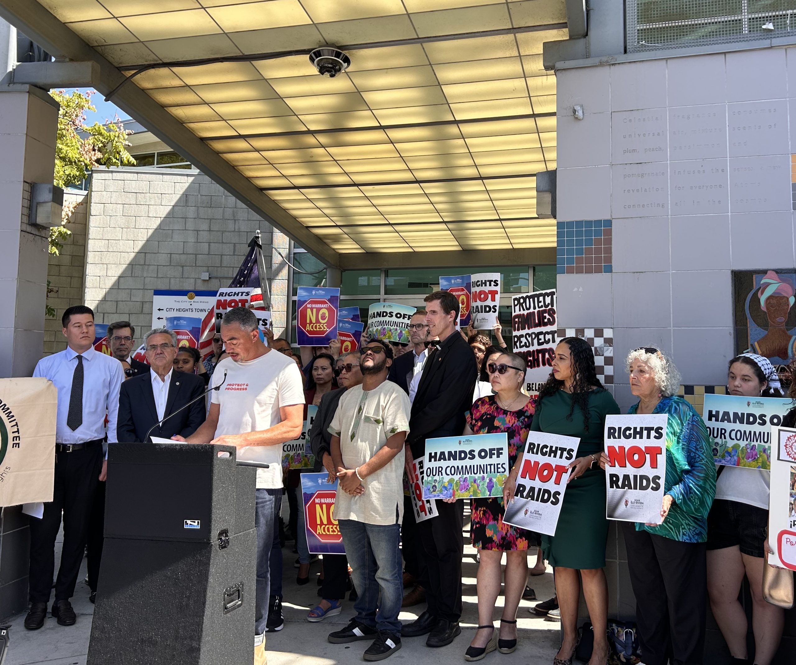 A coalition of politicians at a press conference, with signs that say 'Rights not raids" and "Hands off our communities."