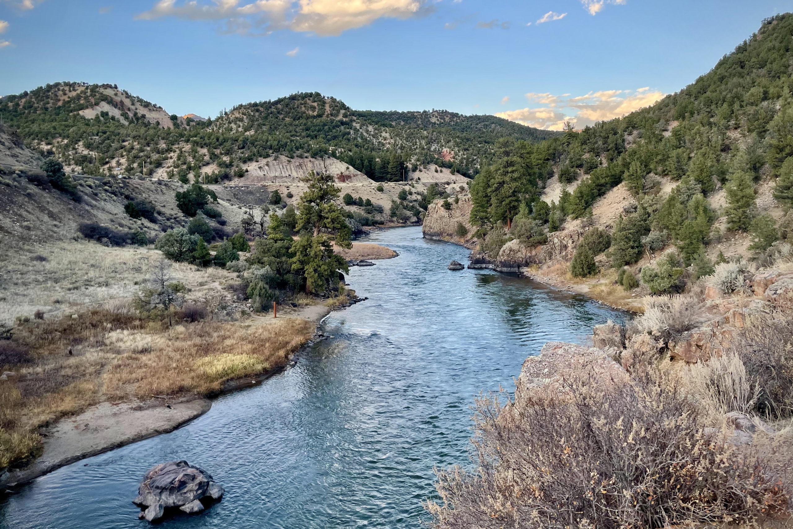 A winding river flows through a valley with rocky banks and tree-covered hills under a cloudy blue sky.