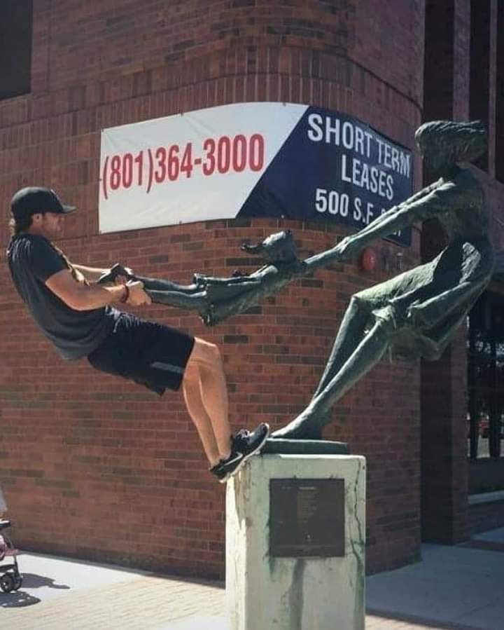A passer-by leans back while holding a bronze girl’s feet, joining a sculpture of a woman swinging a child.