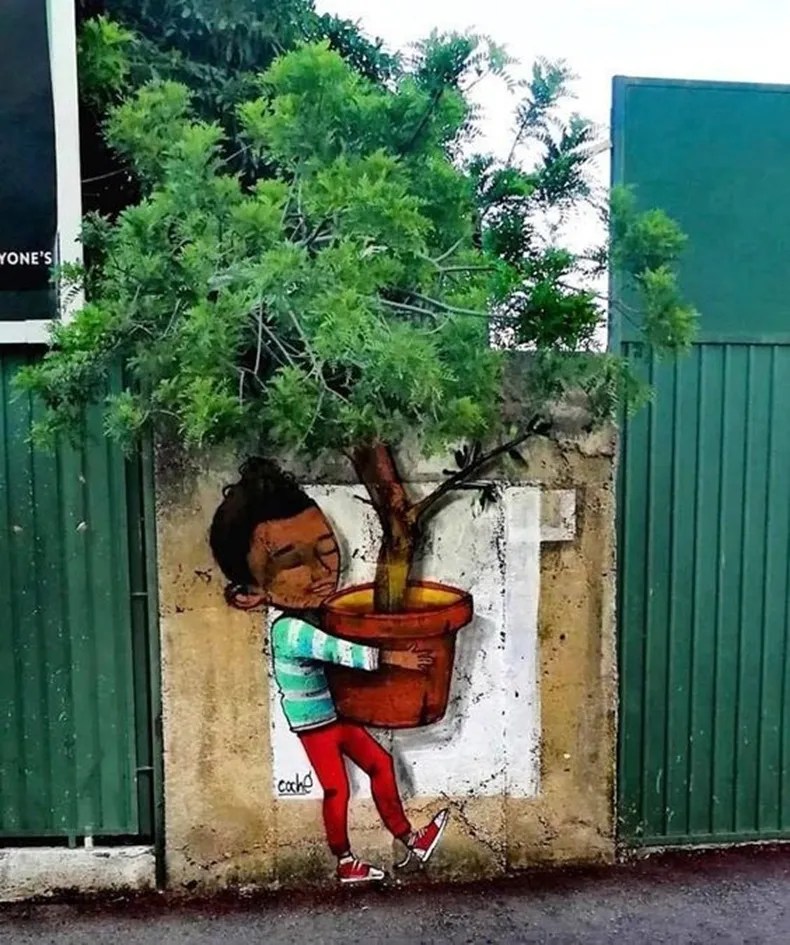A painted child hugs an orange flowerpot on a wall, with a real tree growing behind the wall completing the illusion.