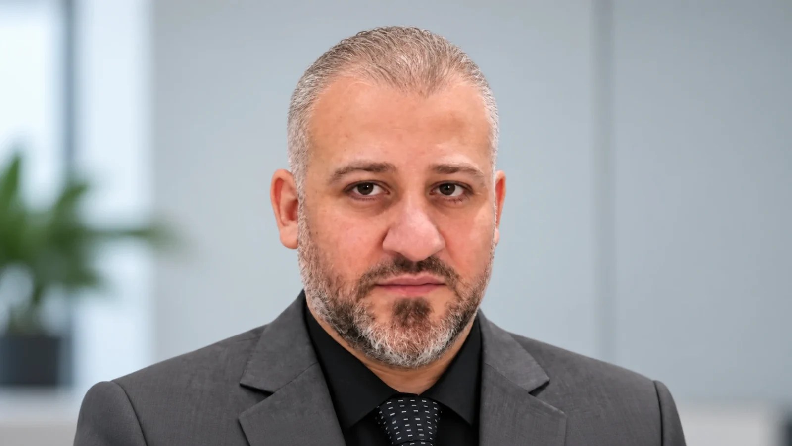 Ayman El-Saleh in a grey suit and black shirt, photographed in a professional indoor setting.