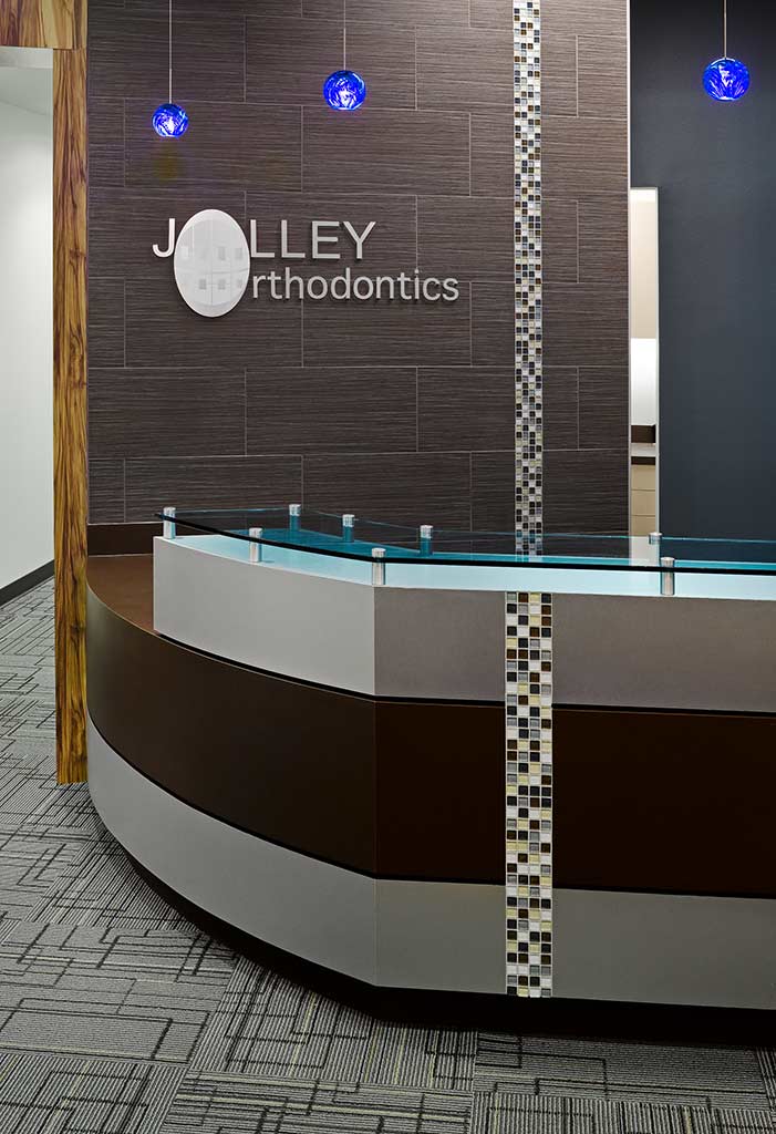 Reception area of Jolley Orthodontics featuring a modern curved desk, decorative wall with the practice name, and blue pendant lights.