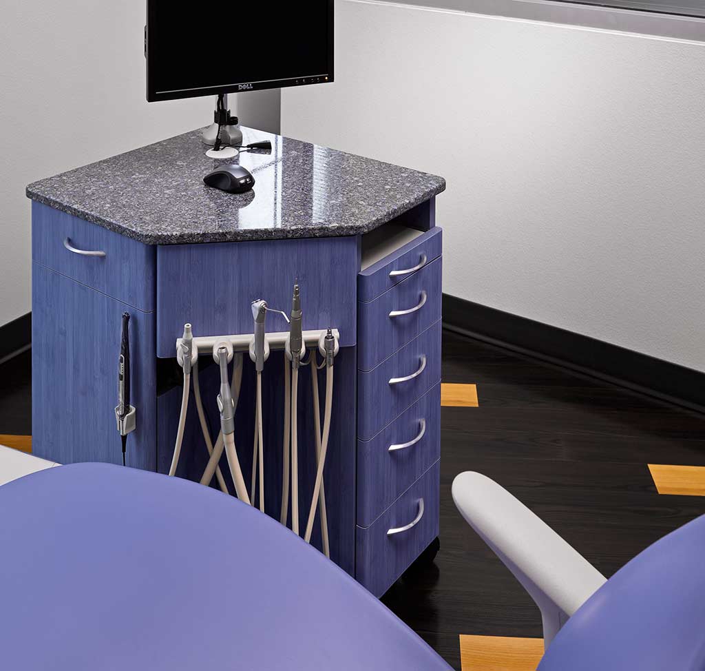 Dental office setup featuring a corner workstation with a monitor, dental tools, and storage drawers, alongside a purple dental chair.