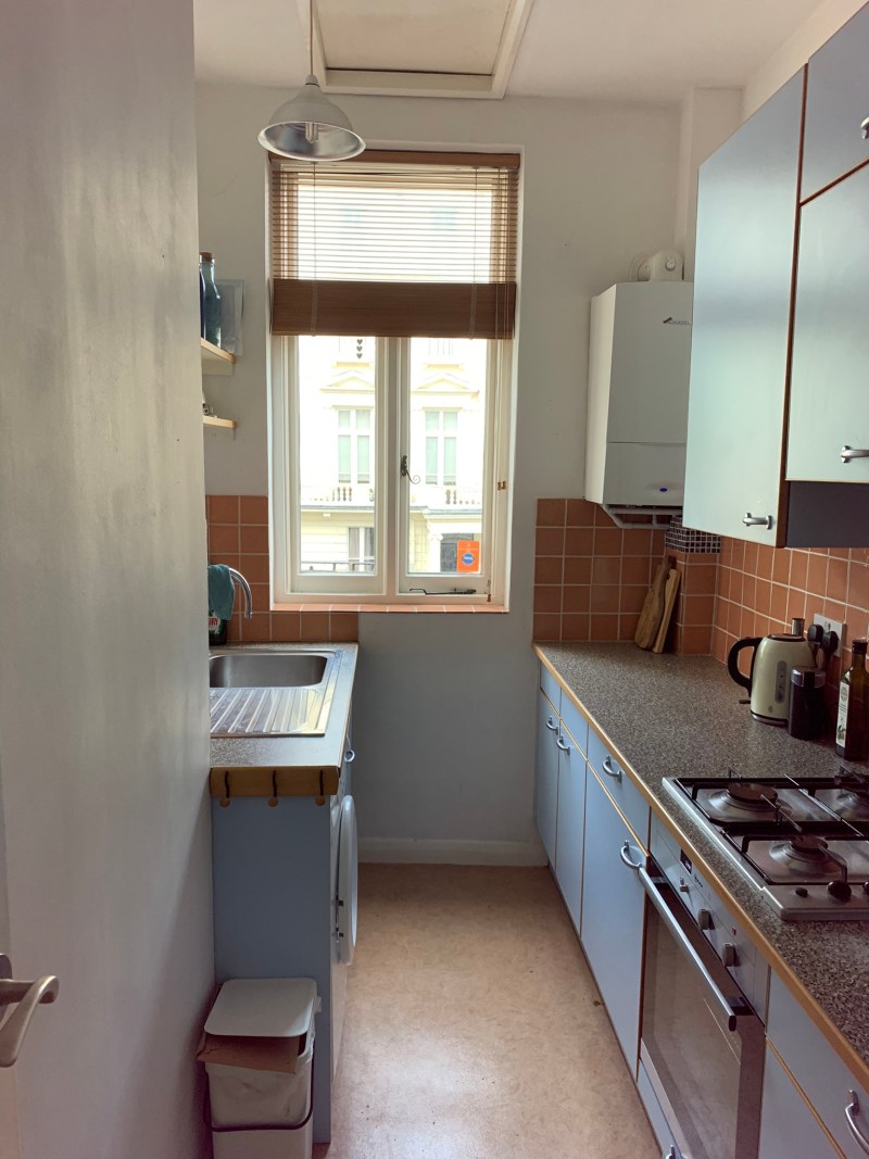A narrow galley kitchen in a period London flat, with pink tiles, plain walls, and a flat dropped ceiling.