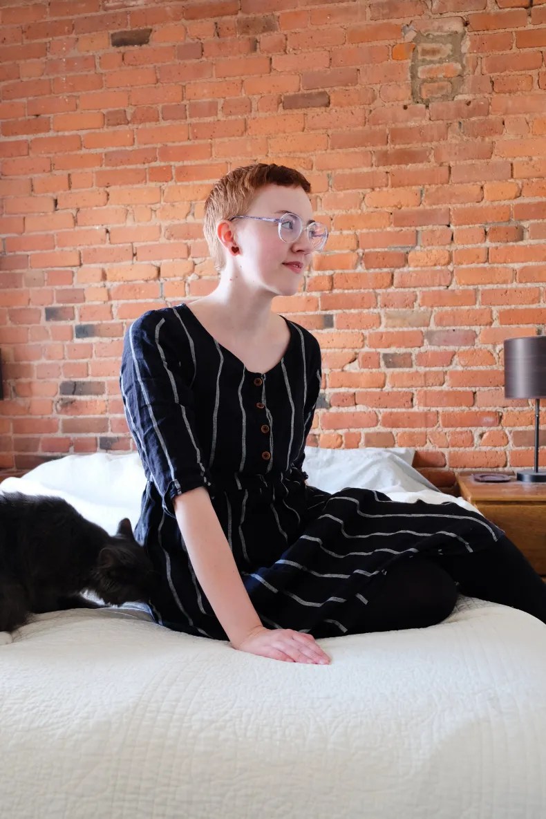 Leah sits on bed with white quilt wearing black pinstripe linen dress