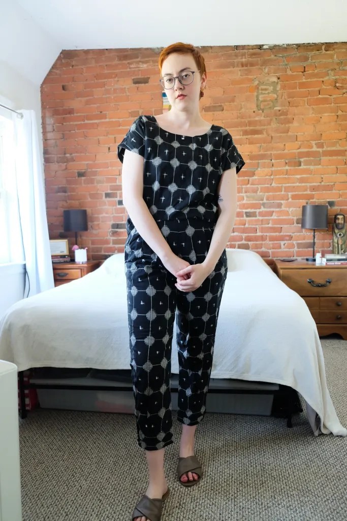 Leah stands in front of bed and brick wall wearing a black patterned jumpsuit and looking serious