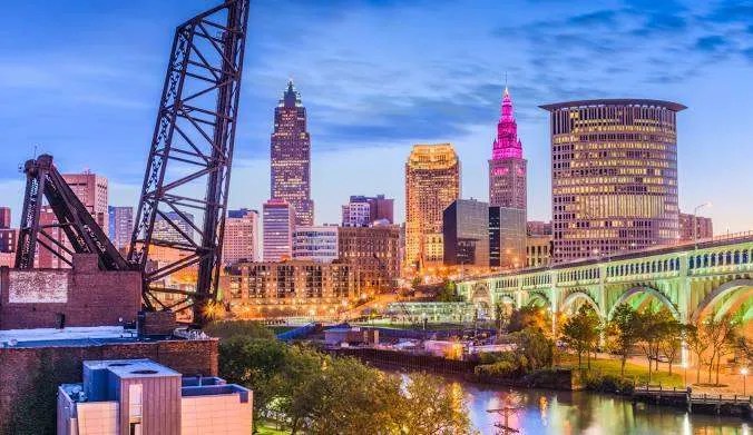 Dusk with colorful lights illuminating tall buildings and a bridge, reflecting in the Cuyahoga River at Cleveland, OH