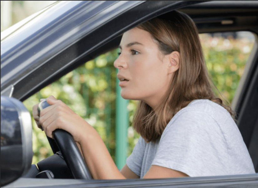 A photograph of a young woman with brown hair sitting in the driver's seat of a black car. She has a worried or intense expression on her face and is gripping the steering wheel tightly with both hands while looking forward out of the window.