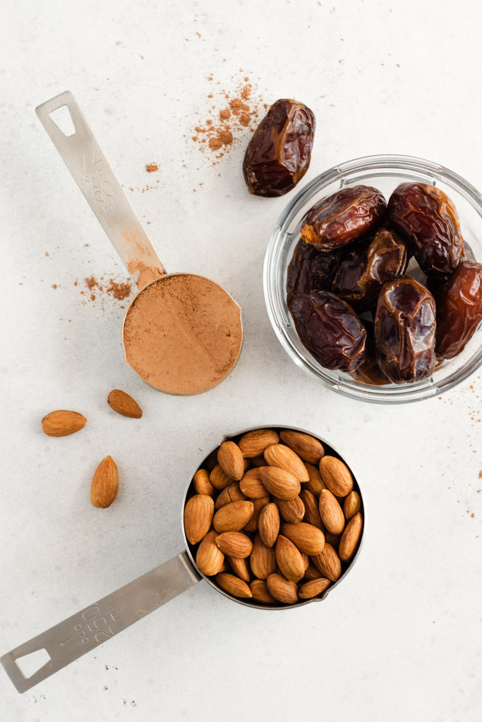 Three round measuring containers, one filled with dates, one almonds and one cacao.