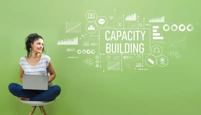 A smiling woman sitting cross-legged on a chair with a laptop, surrounded by illustrated growth charts and icons next to the words "Capacity Building" on a green background.