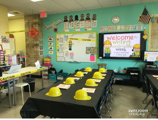 The photo shows a classroom set up for a construction classroom transformation, with a black tablecloth covering the group of desks and hard hats and construction aprons at each seat. A television on the wall says Welcome to Writing Construction Week.