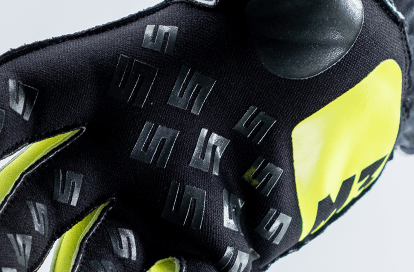 Close-up of a black and neon yellow glove with repeating stylized S logos and textured grip pads on the fingertips.