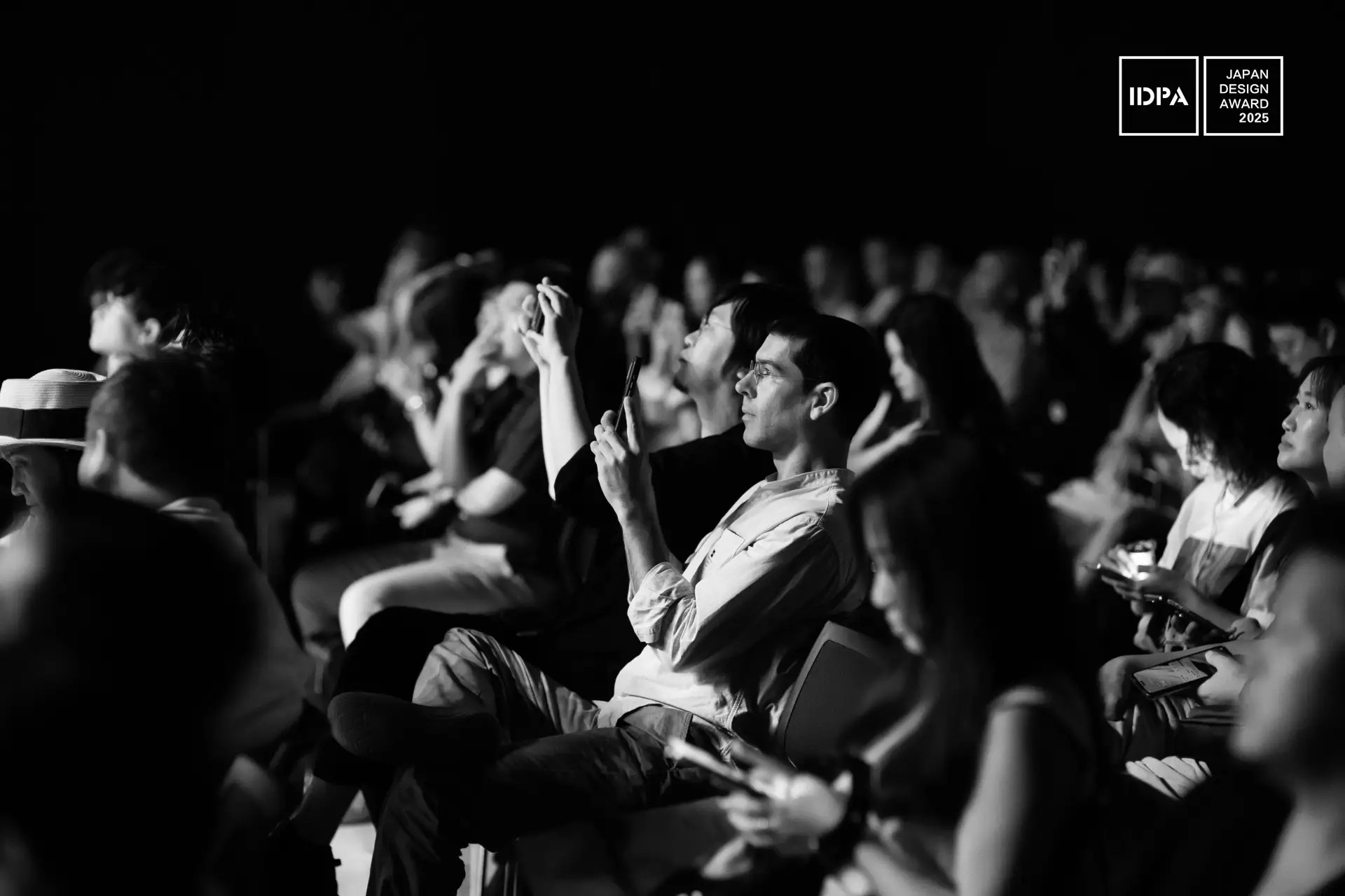Una foto en blanco y negro muestra a un público sentado en un acto, con algunas personas haciendo fotos o usando sus teléfonos. En la esquina superior derecha se ve el logotipo del Japan Design Award 2023.