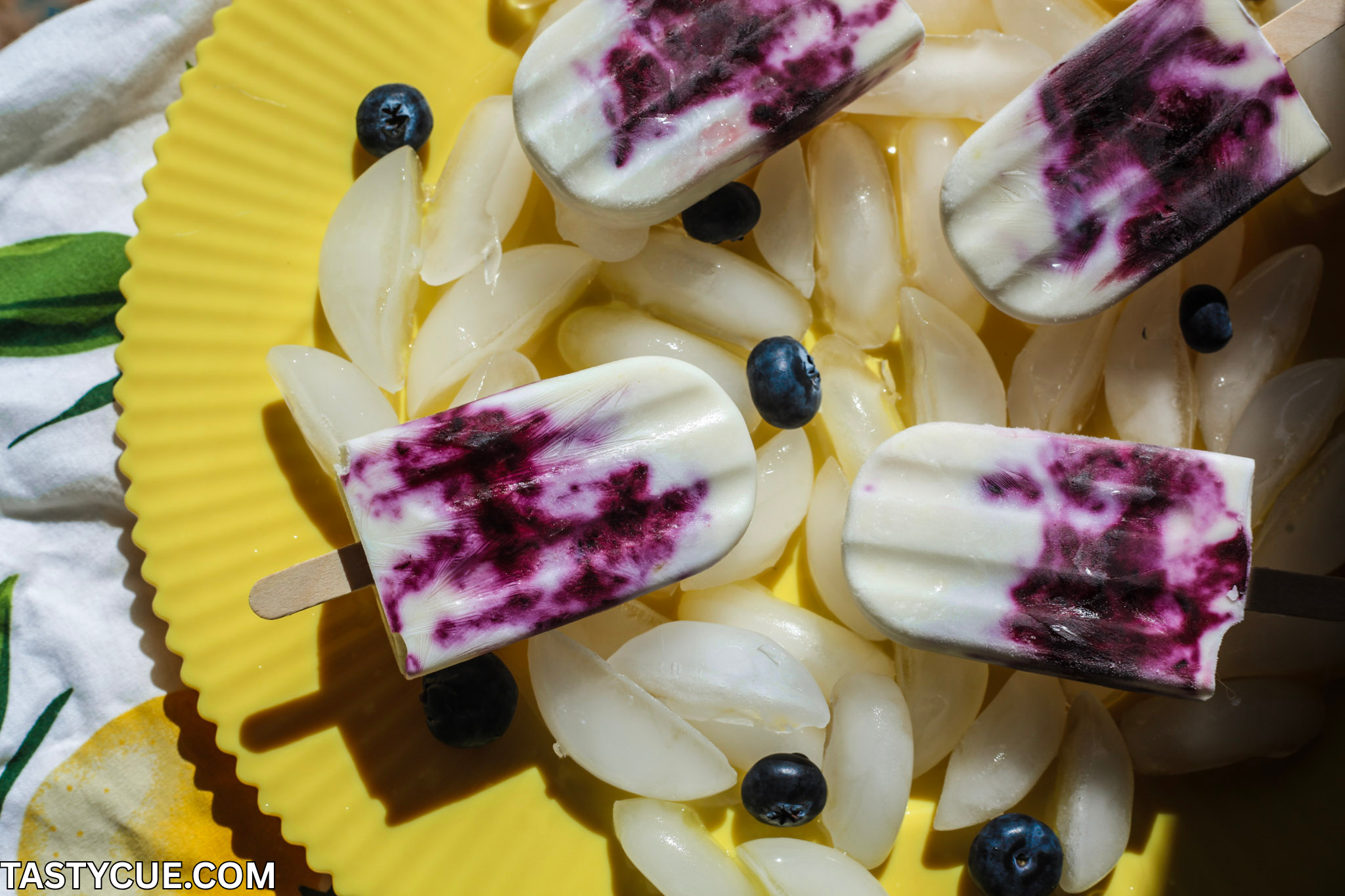 Zitronen-Heidelbeer-Cheesecake-Eis am Stiel auf einem gelben Teller, umgeben von Eiswürfeln und frischen Heidelbeeren.