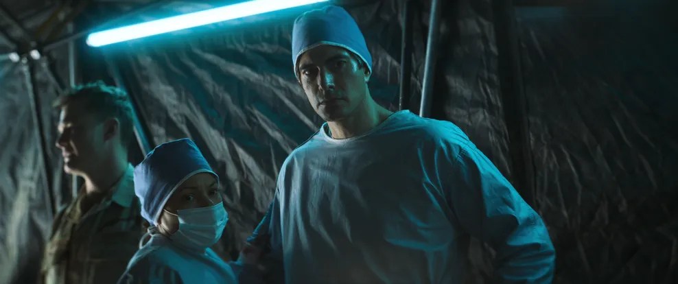 Medical personnel in scrubs and surgical caps standing in a tent with low lighting.