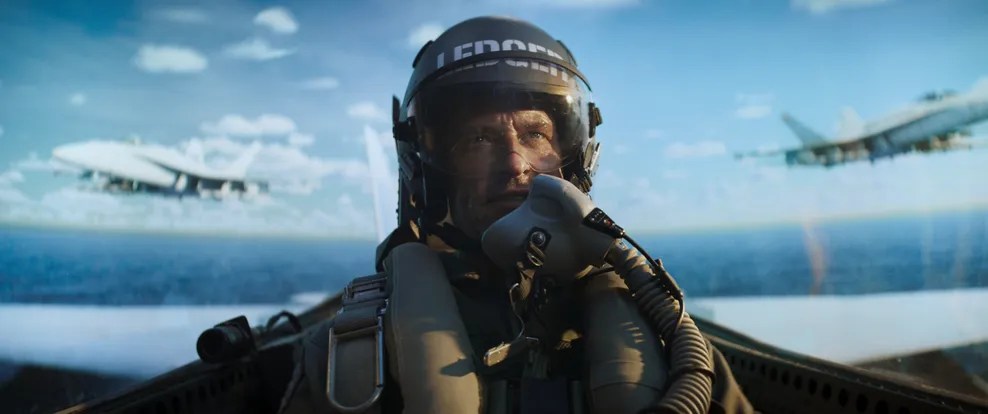 A close-up of a pilot wearing a helmet and oxygen mask inside a fighter jet cockpit, with military aircraft visible in the background and a clear blue sky.
