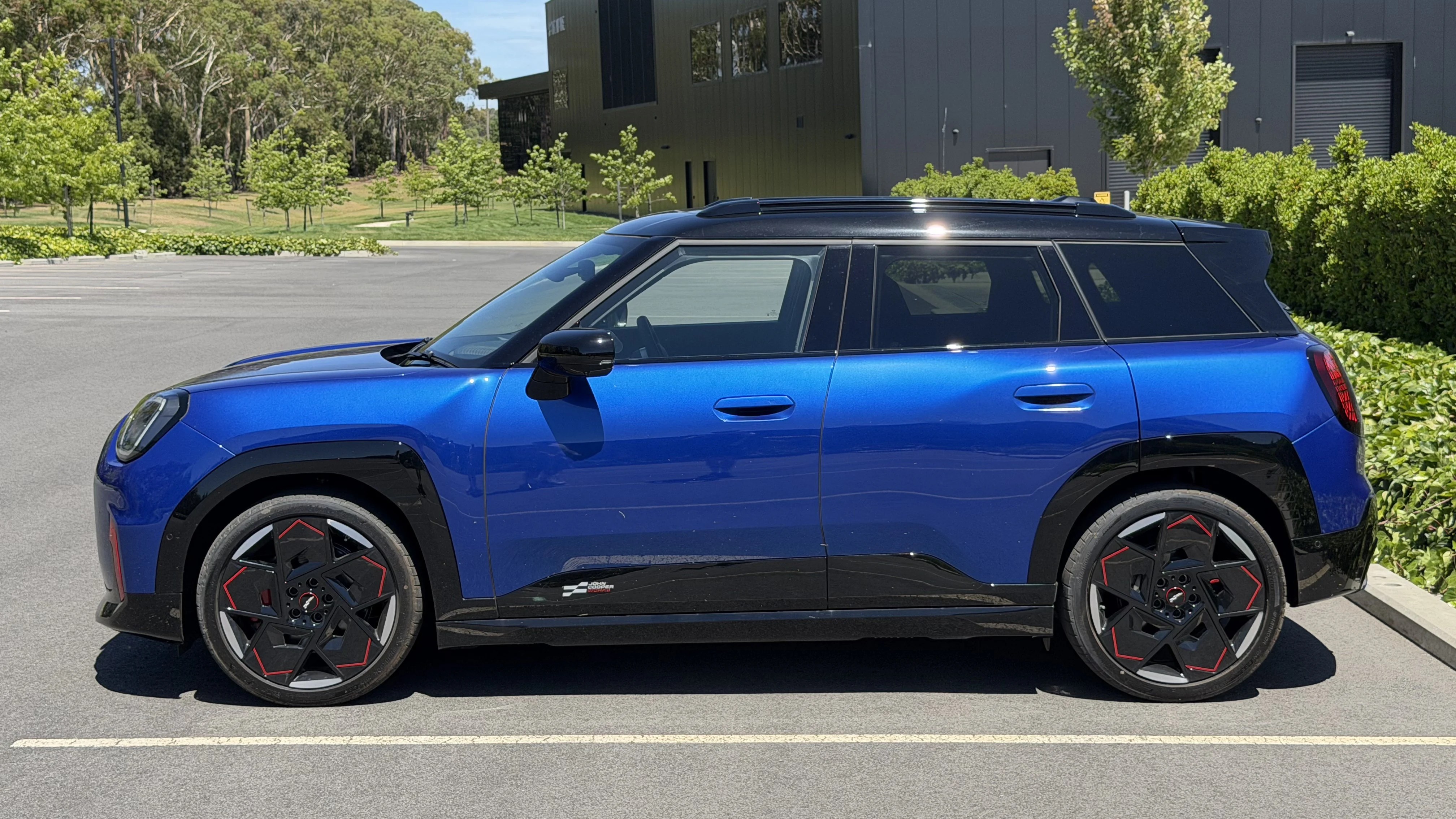 2026 Mini Aceman JCW in blue viewed side-on, black roof and arches, red-accent alloy wheels, parked in outdoor carpark