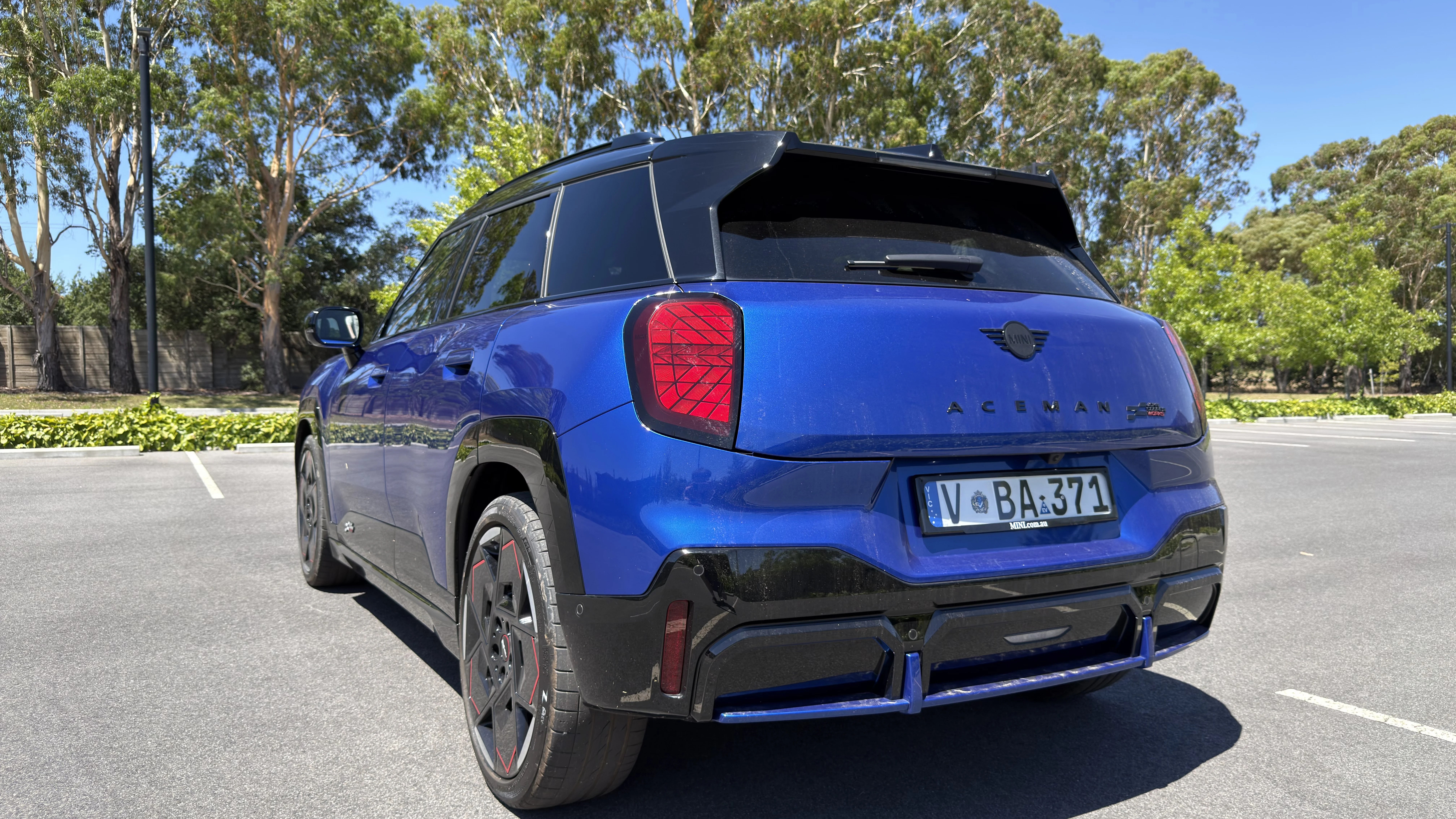 Rear three-quarter of blue 2026 Mini Aceman JCW with black roof, red-accent wheels and rear licence plate in carpark