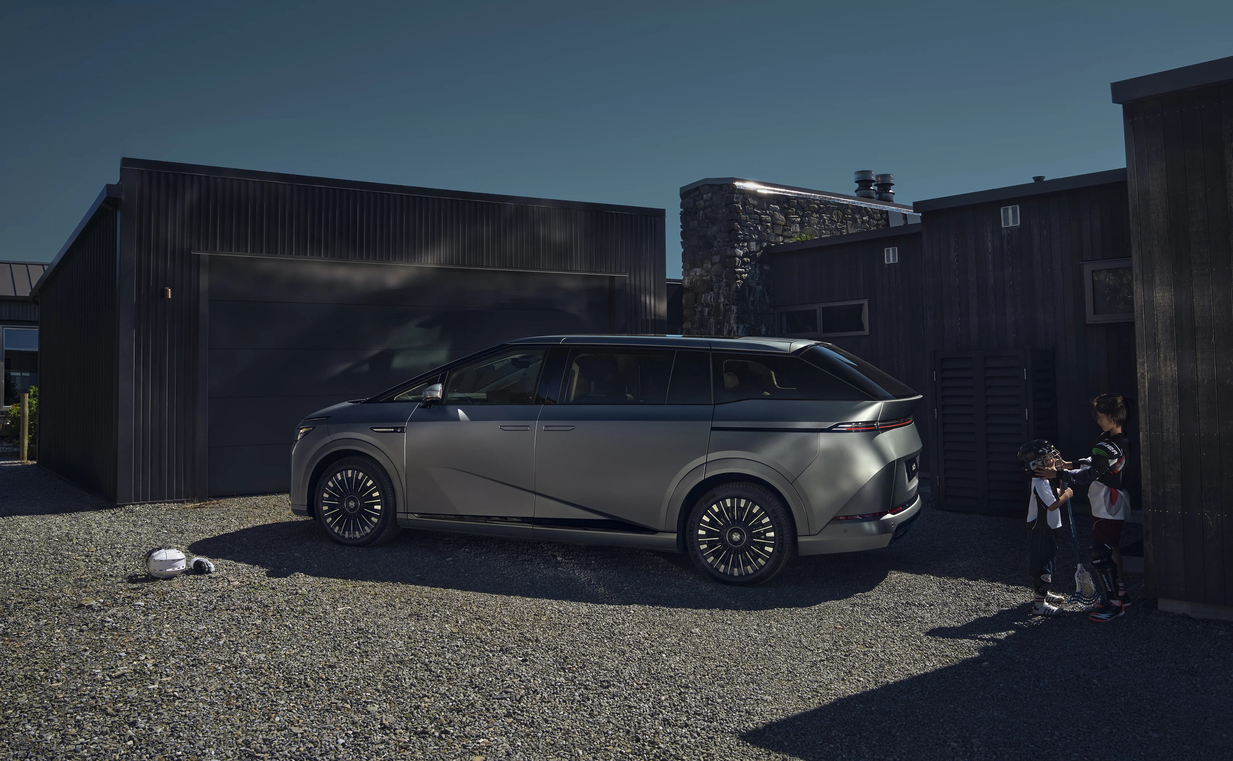 Electric minivan parked in a driveway with two children in protective gear nearby.