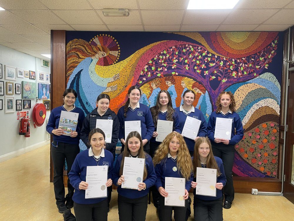 Ten Transition Year students holding their TEFL certificates in a school corridor in front of a large mosaic