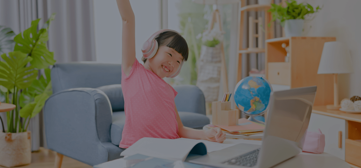 Child raising their hand while on a laptop with a book in front fo her