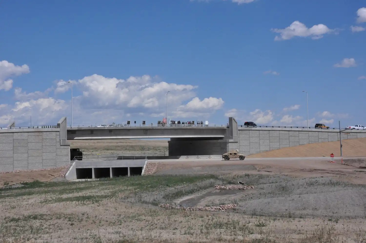 A concrete bridge spans over a dry, grassy area under a blue sky with scattered clouds. Several vehicles are visible on the bridge, and a few construction vehicles and equipment can be seen below the bridge.