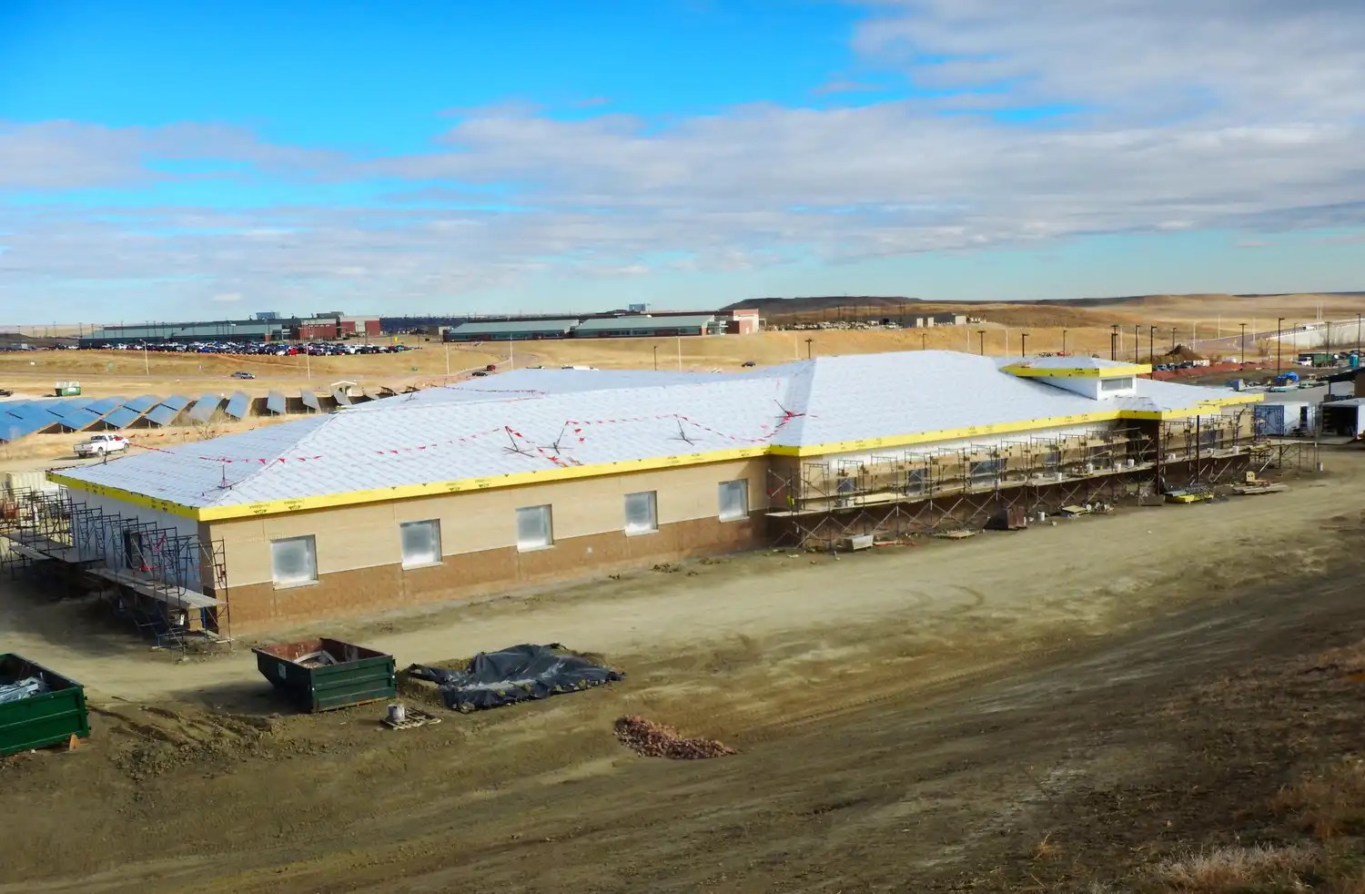 A large building under construction, with scaffolding around its perimeter. The roof is partially installed, and the exterior walls are in progress. The surrounding land is barren and dirt-covered, with construction materials and equipment scattered nearby under a blue sky.