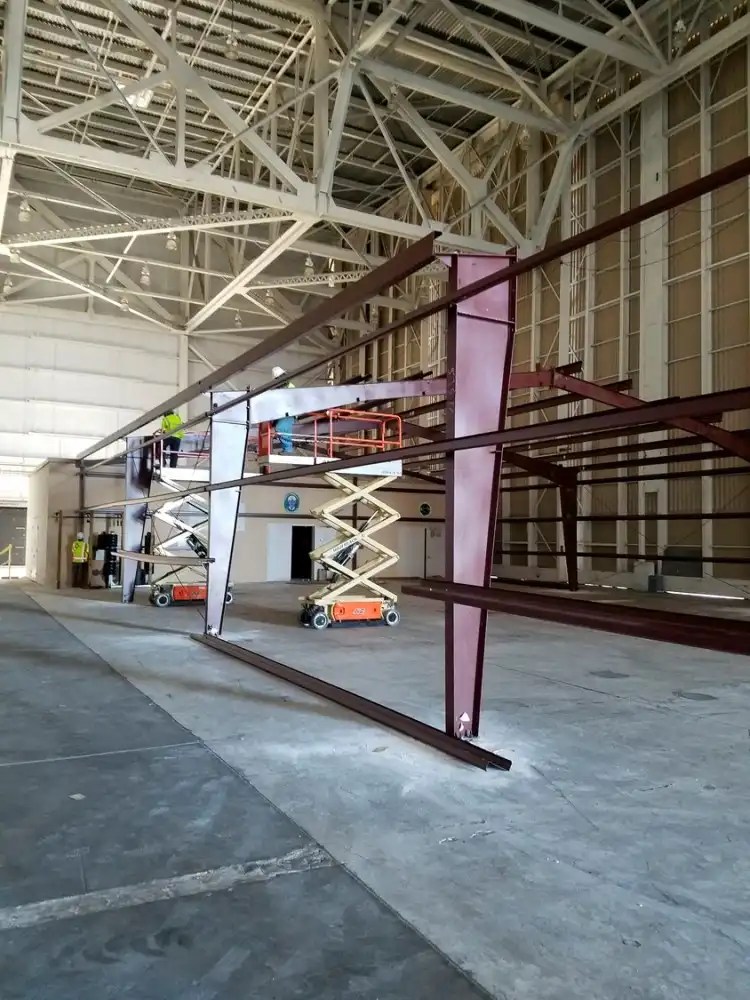 Construction workers are using scissor lifts inside a large warehouse to install a steel frame structure, supporting what appears to be a mezzanine level. The space is expansive, with high ceilings and industrial lighting, and the area is visibly under development.