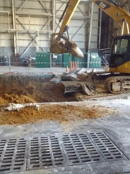 An excavator is operating indoors, digging a large hole in the concrete floor. The surrounding area is marked with caution cones and fencing. There are piles of dirt and concrete rubble around the excavation site. In the background, there are green bins and industrial equipment.