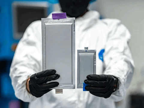 A technician in a cleanroom holding a large conventional battery cell beside a much smaller solid-state cell