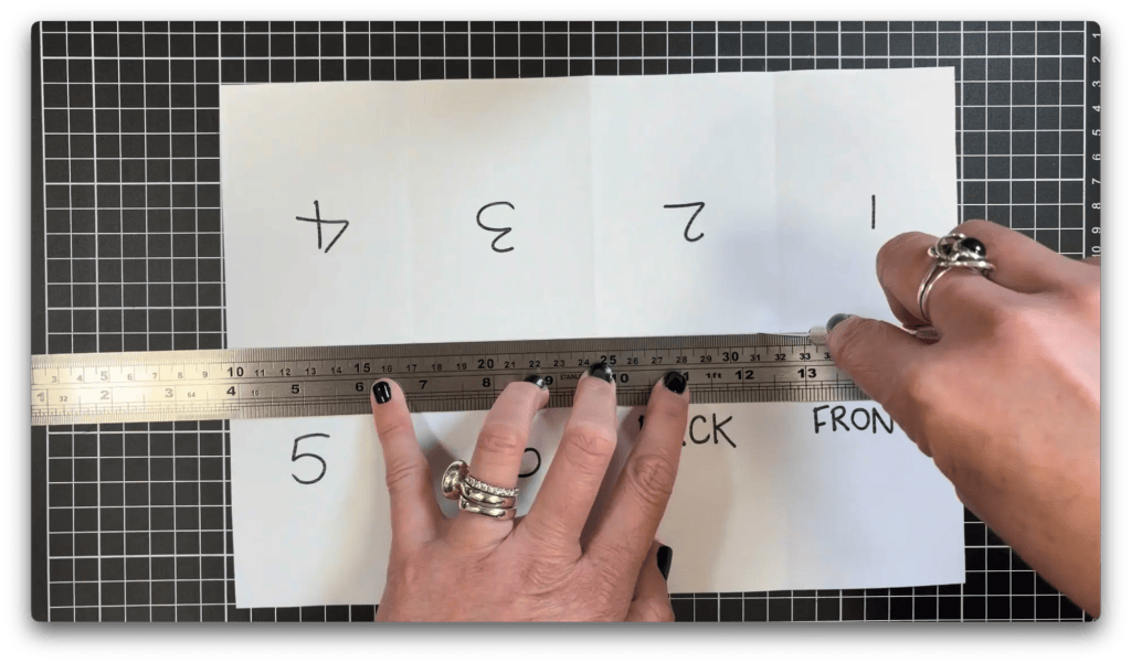 Hands holding a metal ruler and craft knife, cutting a folded paper with numbers 1 to 5 and words "BACK" and "FRONT" on a gridded cutting mat