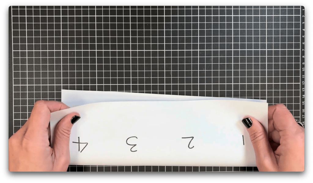 Hands folding a paper numbered 1 to 4 on a black grid cutting mat