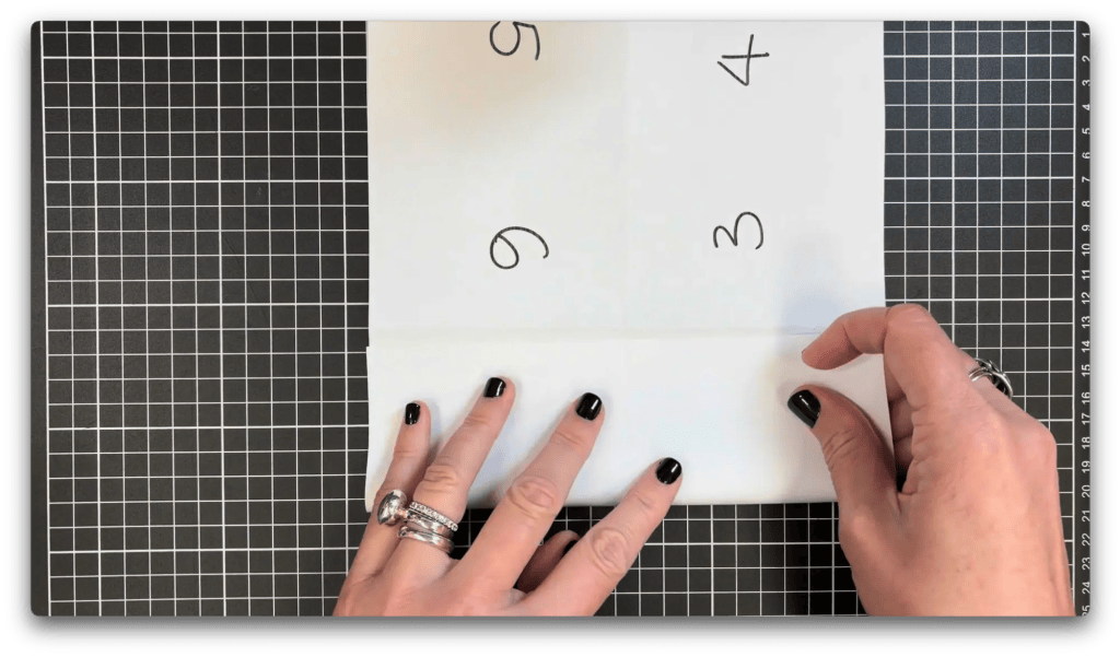 Two hands folding a white paper with numbers 3, 4, 5, and 9 written on it over a black and white grid cutting mat