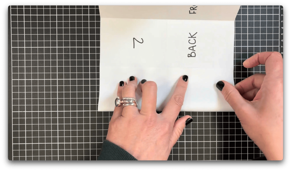 Hands with black nail polish holding folded paper with handwritten words 'BACK' and number '2' on a black and white grid cutting mat