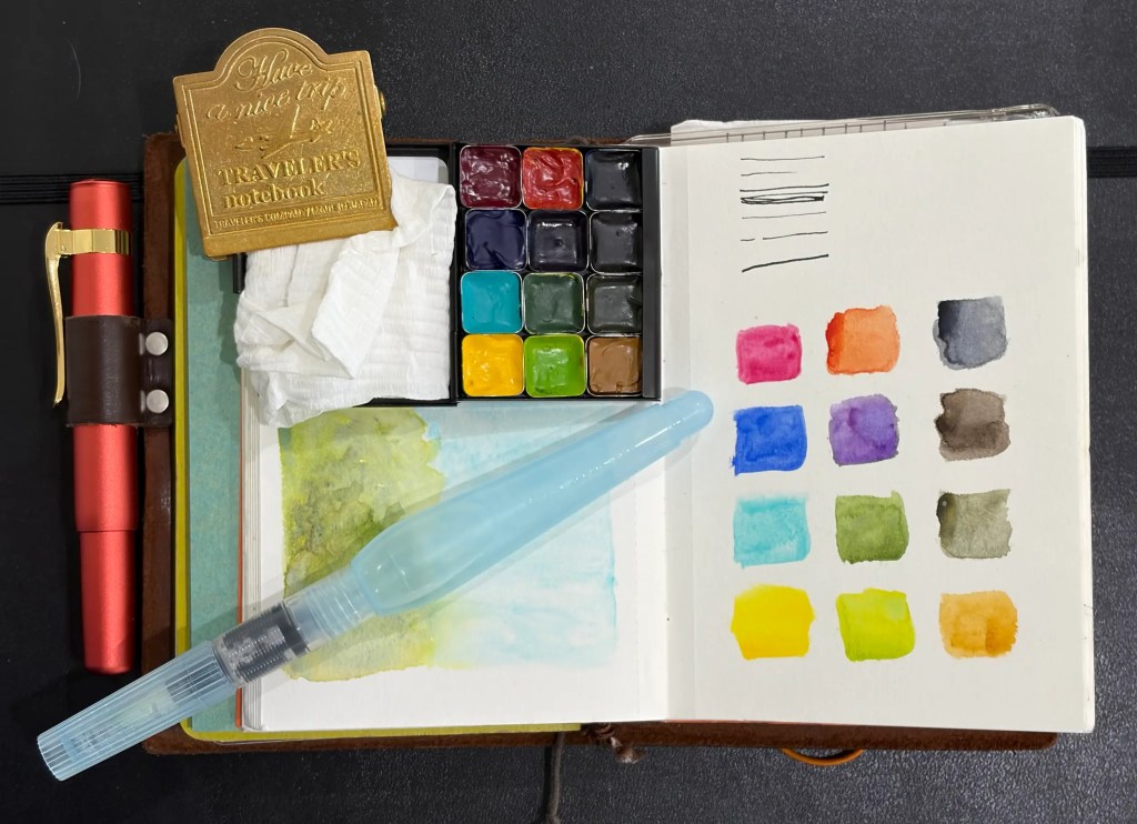 Open watercolor palette with various color pans, a brown traveler's notebook with a gold metal clip embossed with 'Have a nice trip TRAVELER'S notebook', a red pen in a leather holder, a water brush, and a notebook page showing watercolor swatches and ink lines