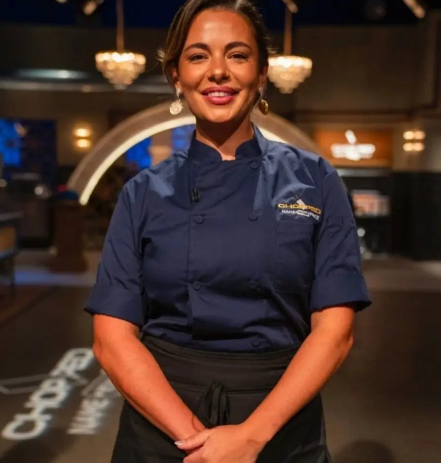 Professional studio portrait of a smiling female executive chef in a navy blue chef uniform and black apron inside a restaurant setting.