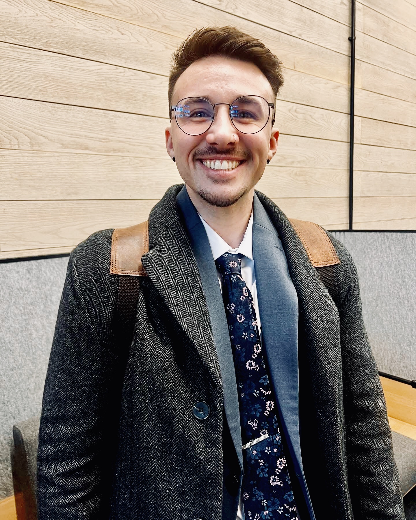 An individual with round glasses, a short cropped hair do who is smiling wearing a dark grey jacket and a patterned navy tie in front of some wooden panels.