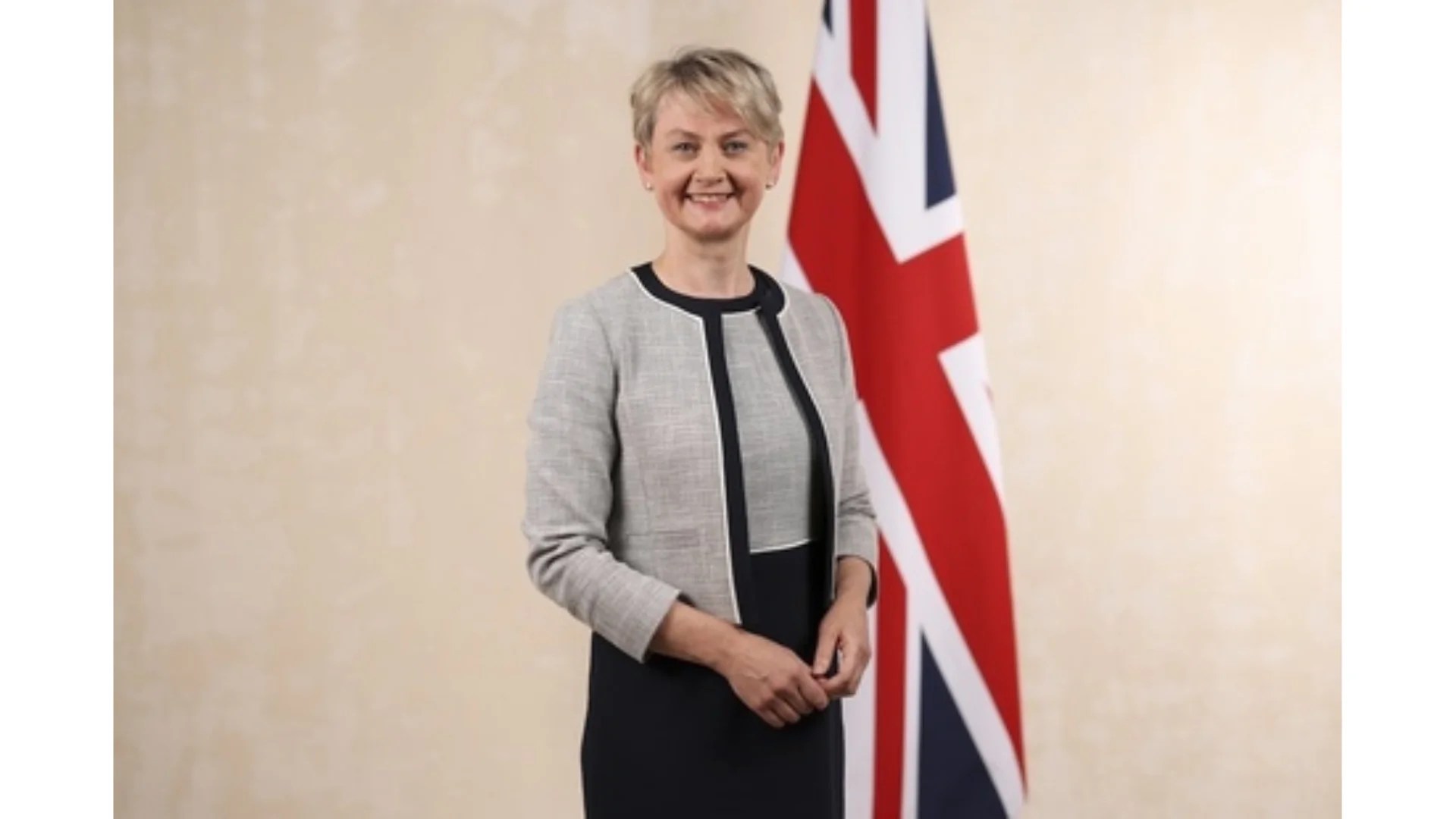 Image of Home Secretary Yvette Cooper standing in front of a Union Jack flag against a beige background.