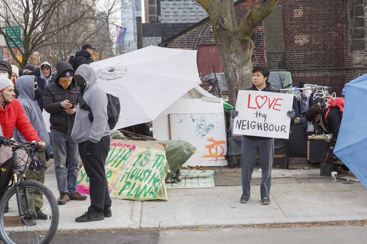 A person with a sign that reads "Love Thy Neighbour" stands outside the encampment at St. Stephen in-the-Fields.
