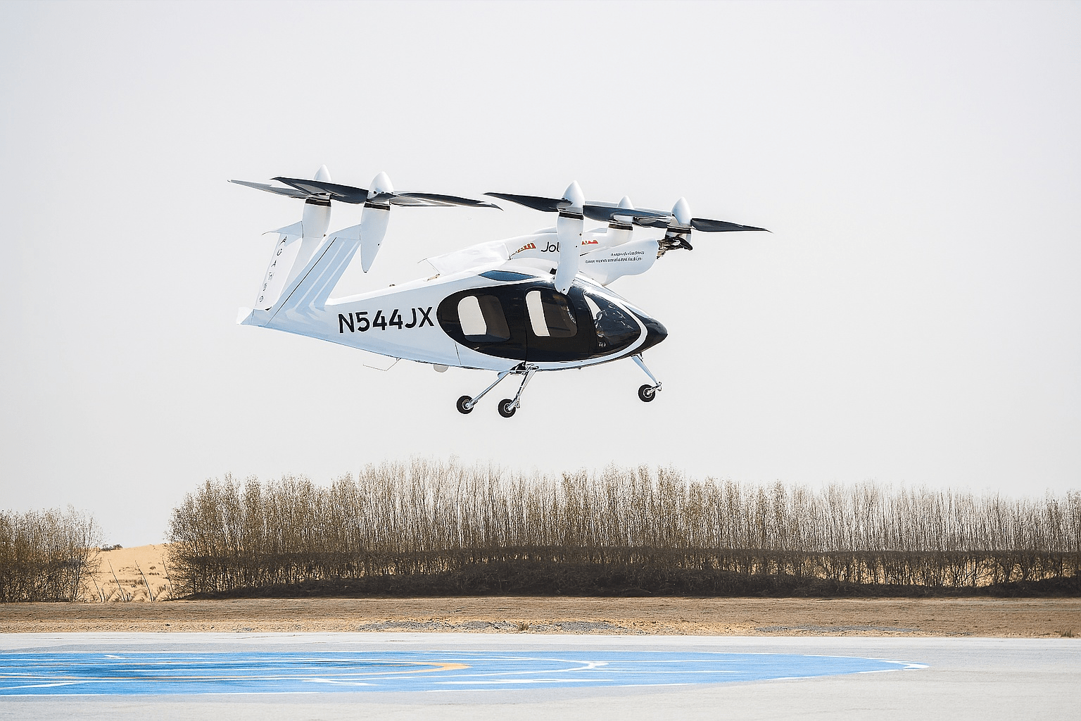Joby’s electric air taxi in mid-flight during a test in Dubai, with six rotors visible above its white and black fuselage, against a desert backdrop and helipad below.