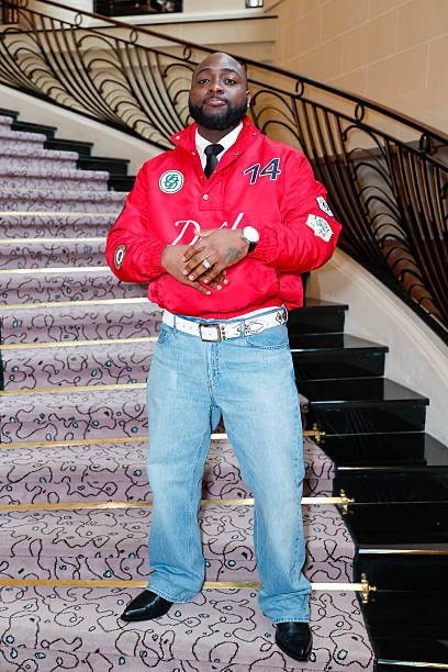 A man stands confidently on a staircase wearing a red jacket, jeans, and a white belt, accessorised with a watch.