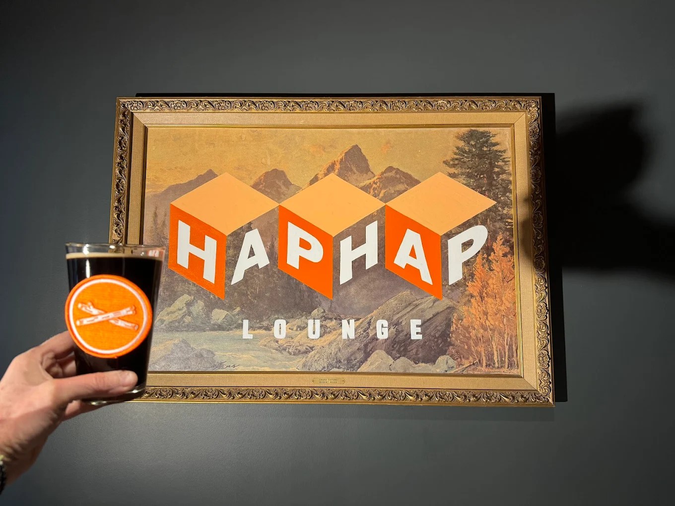 Hand holding a dark beer in front of the Hap Hap Lounge sign in Boise.