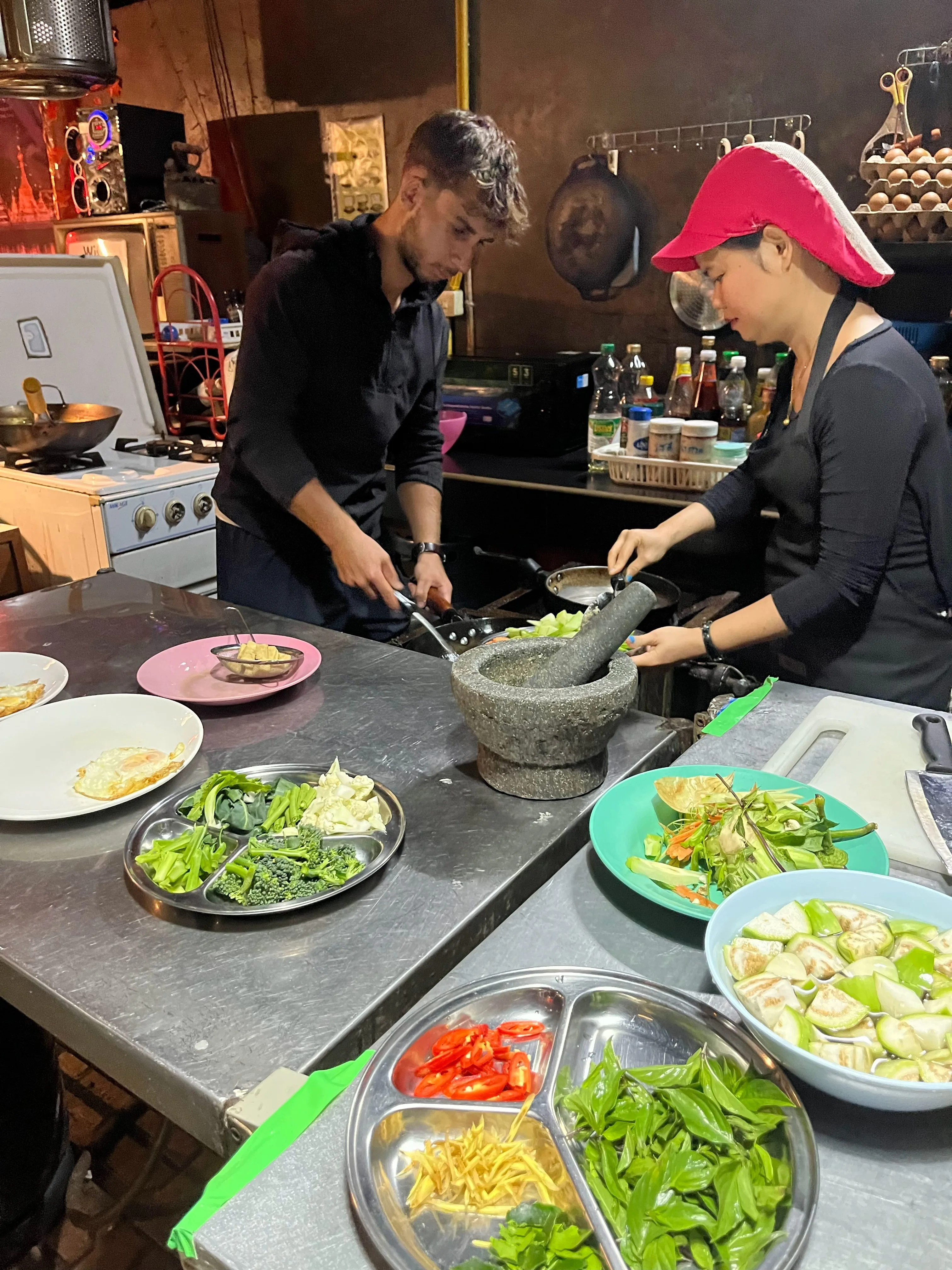 me cooking next to the thai cook in the kitchen at crossroads house in mae hong son, with fresh vegetables and a mortar and pestle on the counter