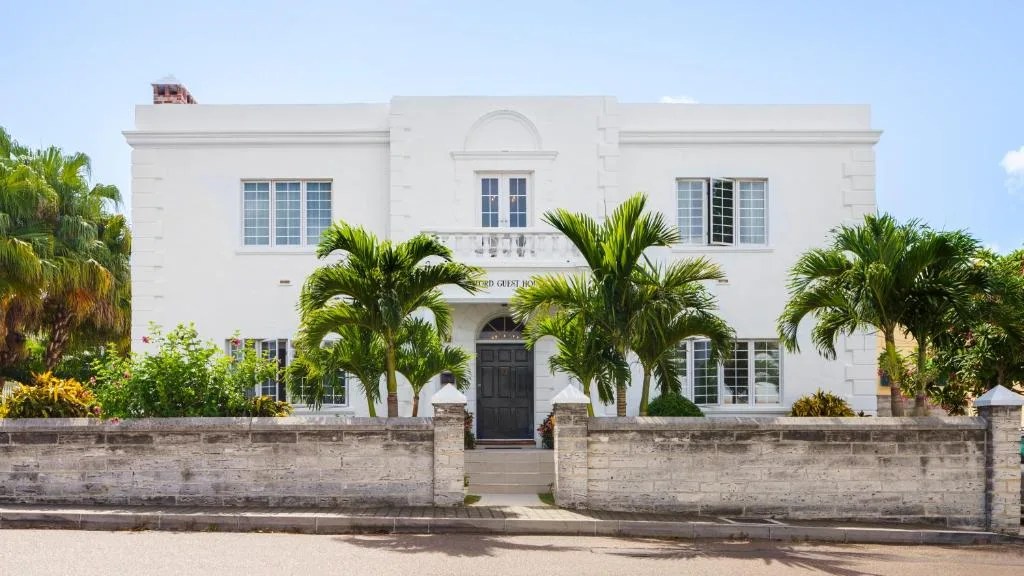 white two-story oxford house guesthouse behind a stone wall and palm trees in hamilton, bermuda
