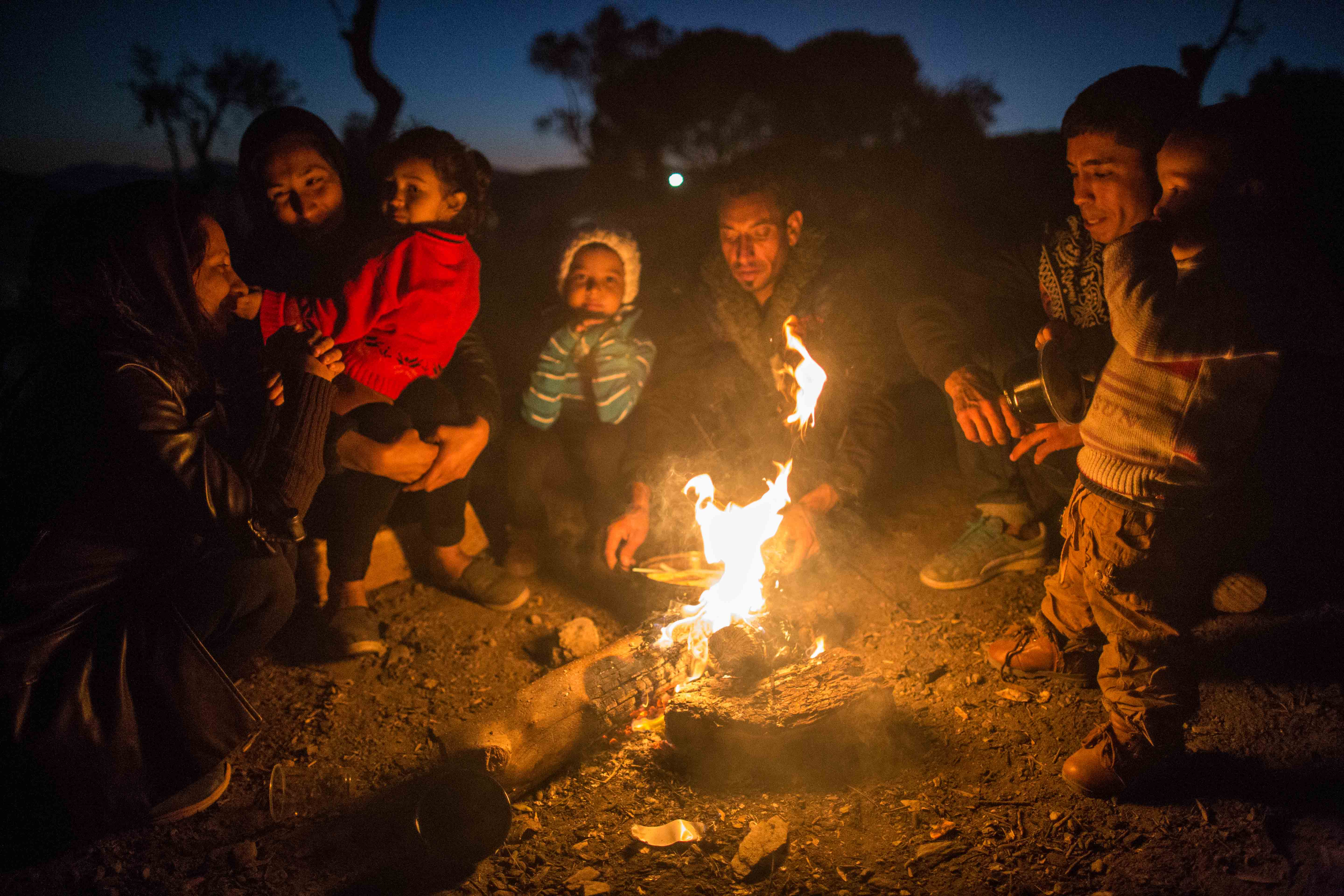 Dusk at Moria Camp. Families outside the camp fence huddle around open fires in olive orchard.