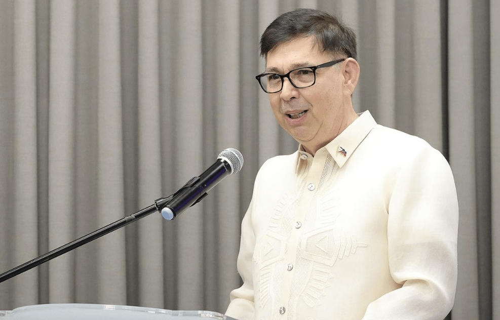 A man in a traditional Barong Tagalog, wearing glasses, stands at a podium speaking into a microphone against a backdrop of gray curtains.