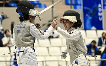 Two female fencers celebrate a match victory, fist bumping each other while wearing protective gear and fencing masks.