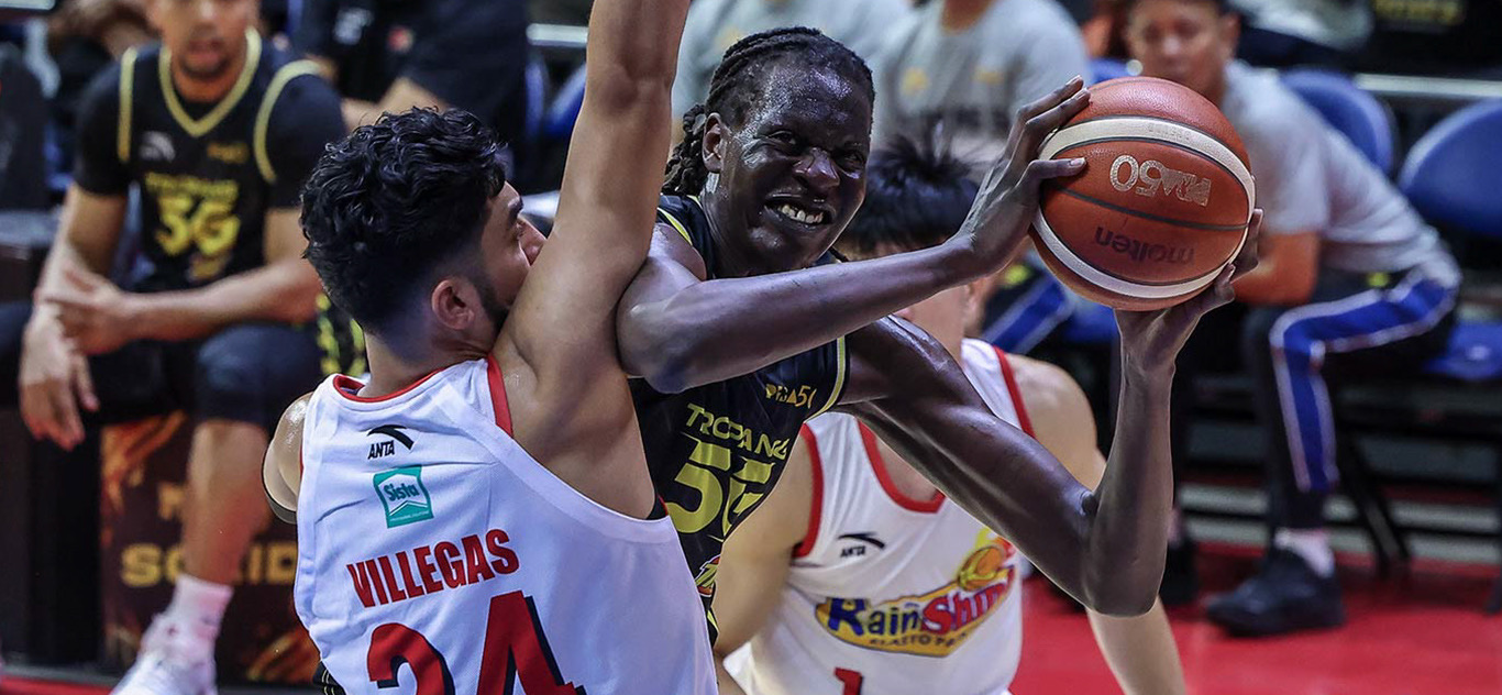 A basketball player in a black jersey struggles to maintain possession of the ball while being closely guarded by an opponent in a white jersey. The scene highlights the intensity of a competitive game.