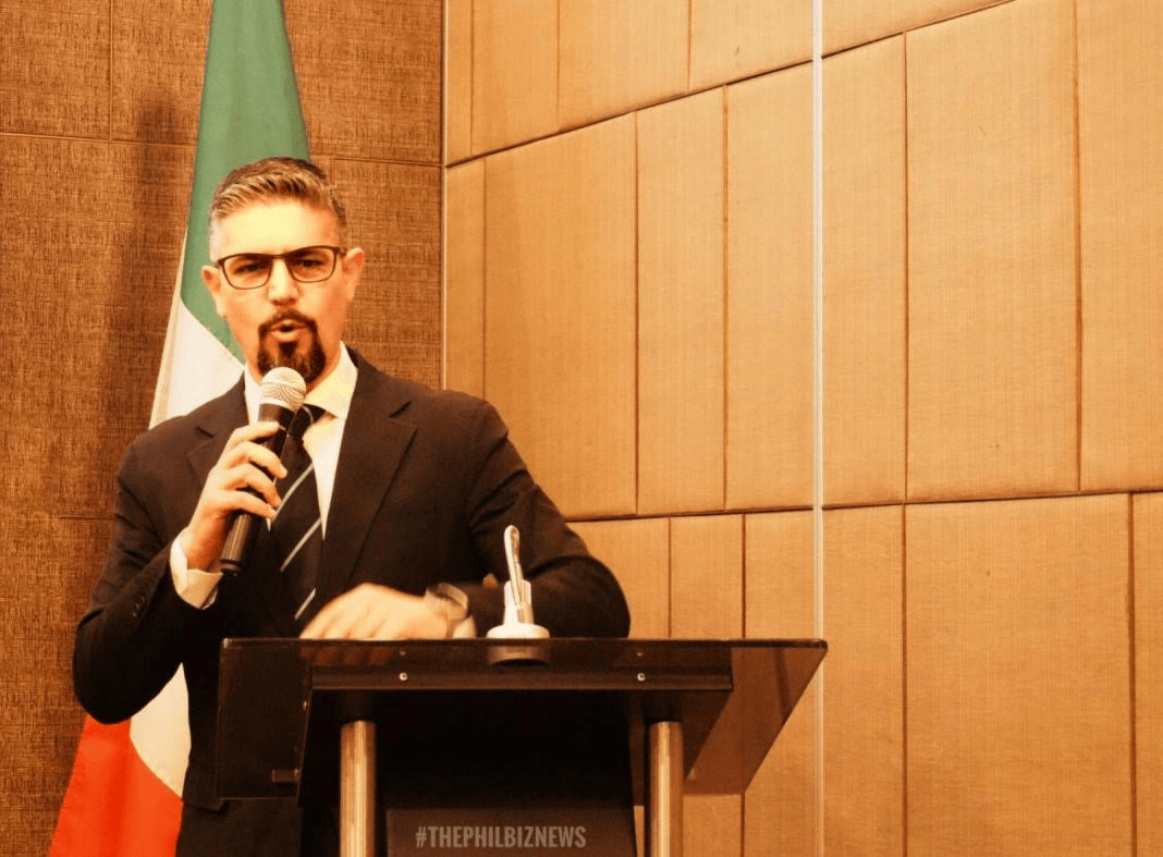 A man in a suit and glasses speaking at a podium with a microphone, with the Italian flag in the background.